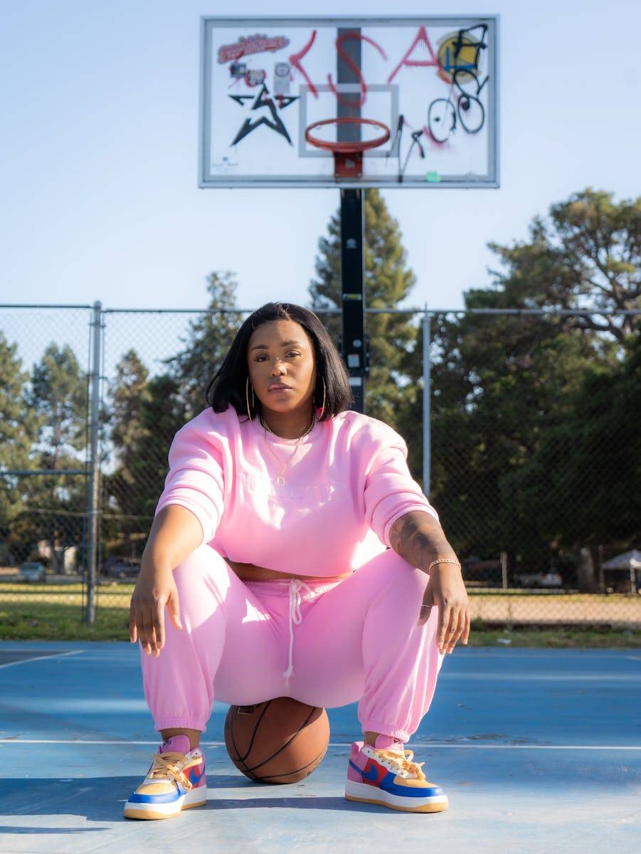 A woman wearing a pink tracksuit poses confidently on a basketball court outdoors.