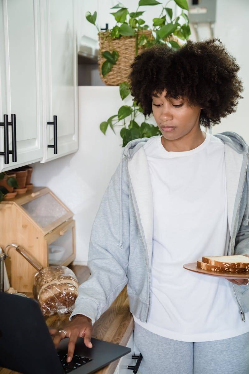Young woman multitasking in kitchen with laptop and breakfast in hand