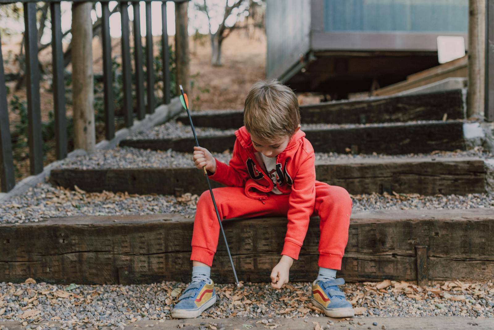 Child in red tracksuit holding arrow, sitting on wooden steps outdoors.