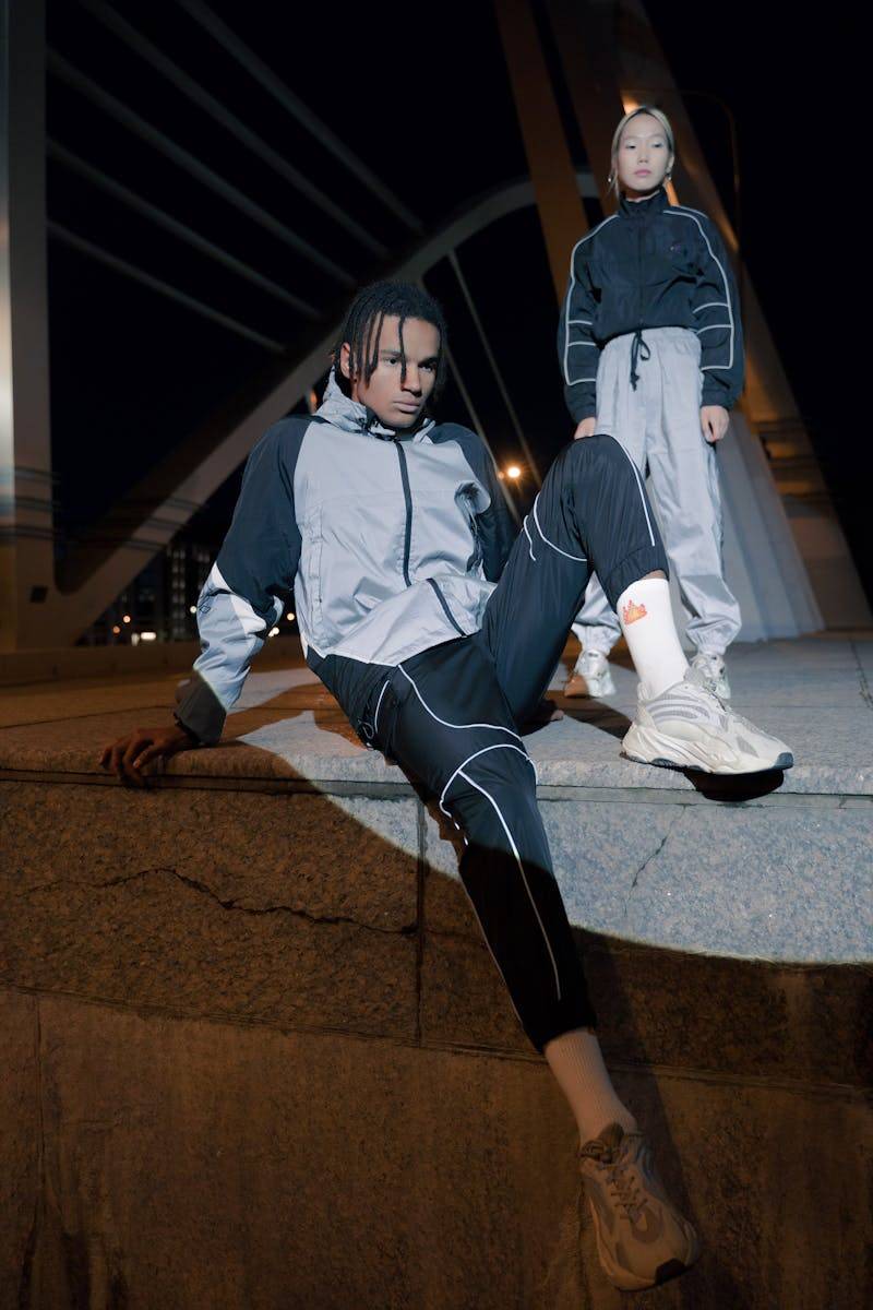 Two individuals in sporty attire pose under a city bridge at night, showcasing modern street style.