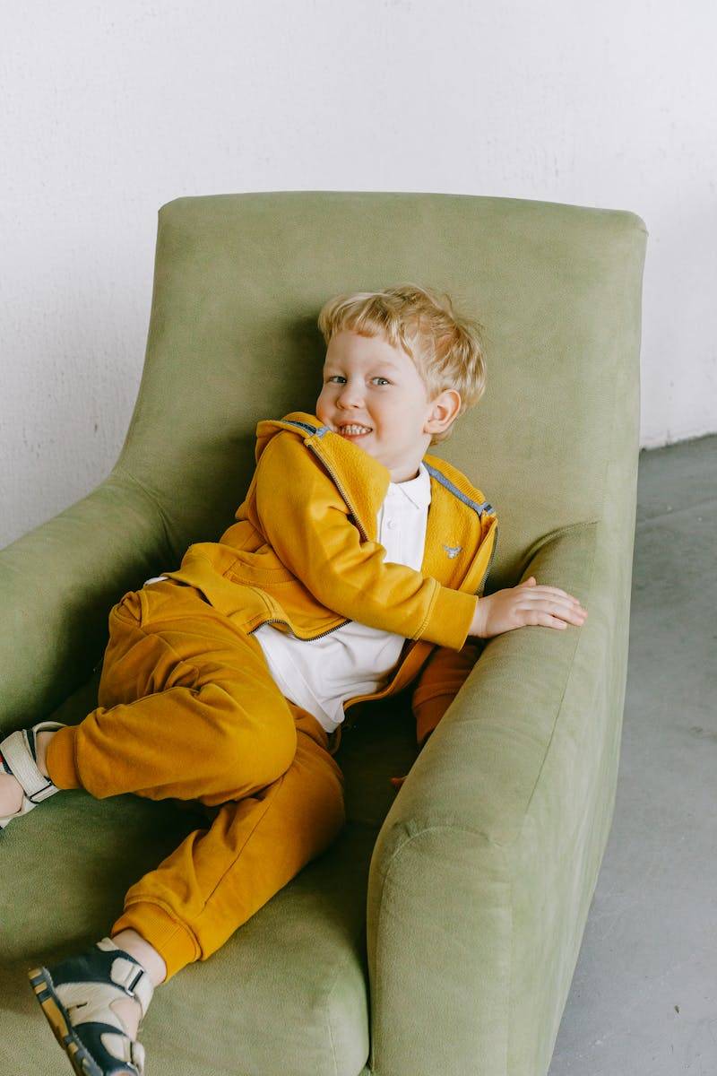 Young boy in a yellow tracksuit lounging happily in a green armchair indoors.
